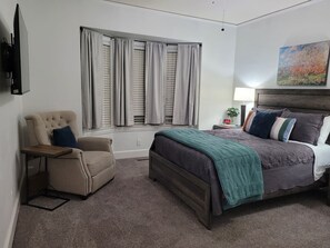 Ein Schlafzimmer mit Bett, Sessel, Nachttisch, Lampe und einem Fenster mit Vorhängen.
