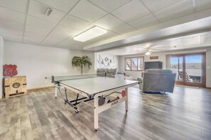 Ein Zimmer mit einem Pongtisch, einer Couch, einem Sessel, einer Pflanze und Blick auf einen See durch eine Glastür.