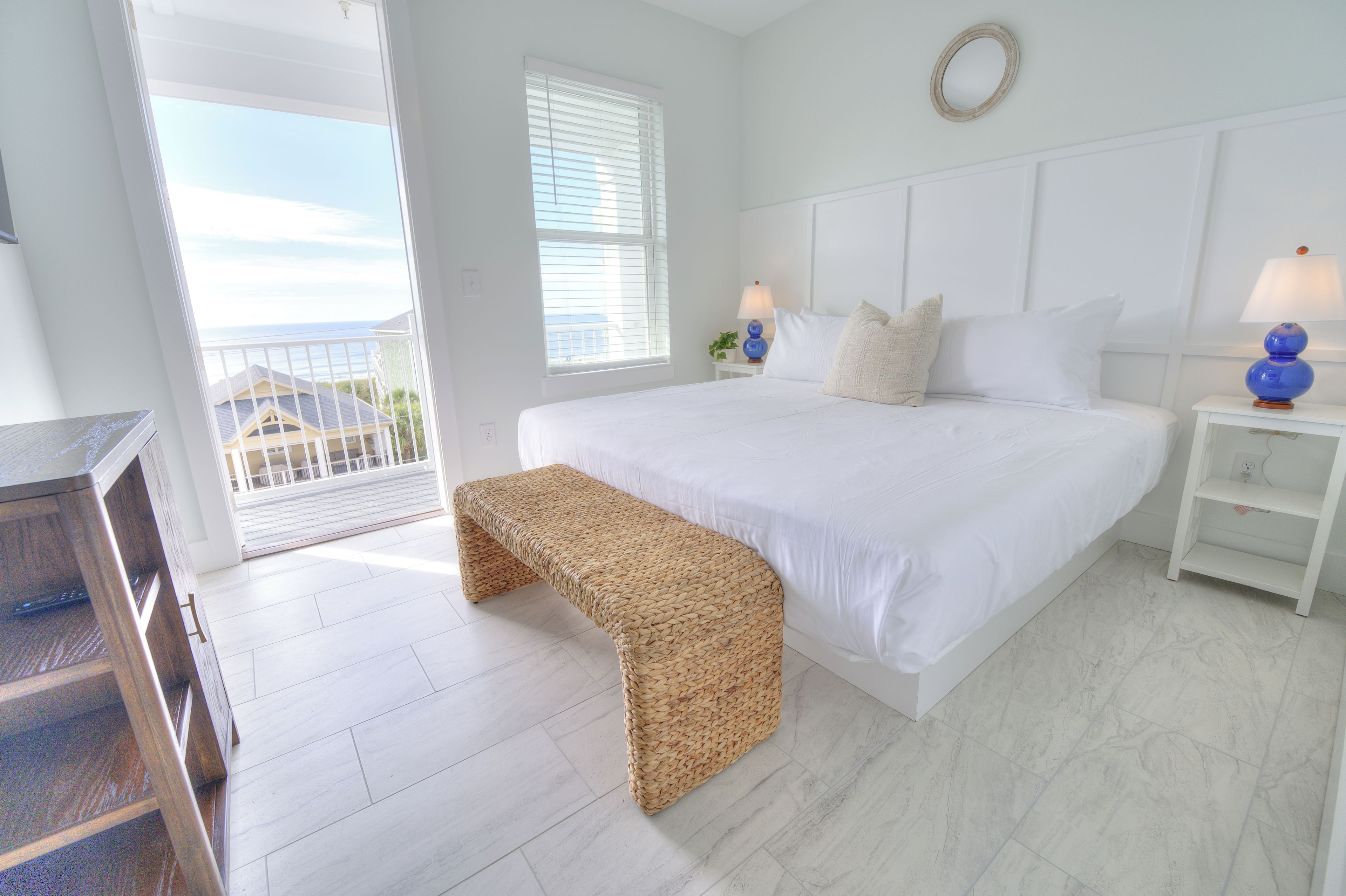 A bedroom with a large bed, a bedside table with a lamp, a bench, and a view of the sea through the window.