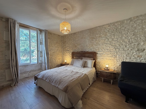 Un dormitorio con pared de piedra, cama con cabecera de madera, dos mesitas de noche con lámparas, una ventana con cortinas y un sillón oscuro.