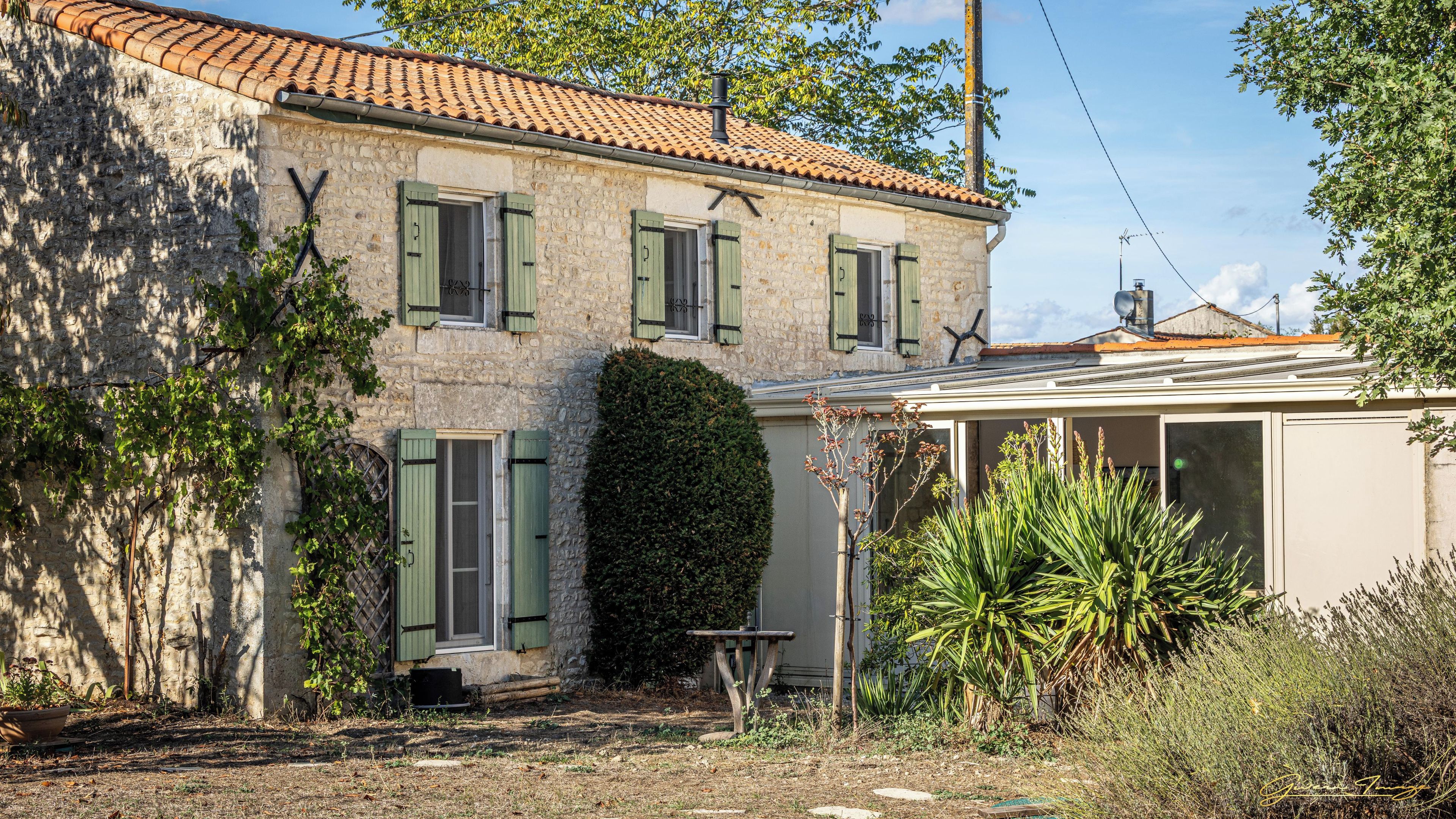 A stone building with a tiled roof, green shutters, and a small garden area with plants and a bench.