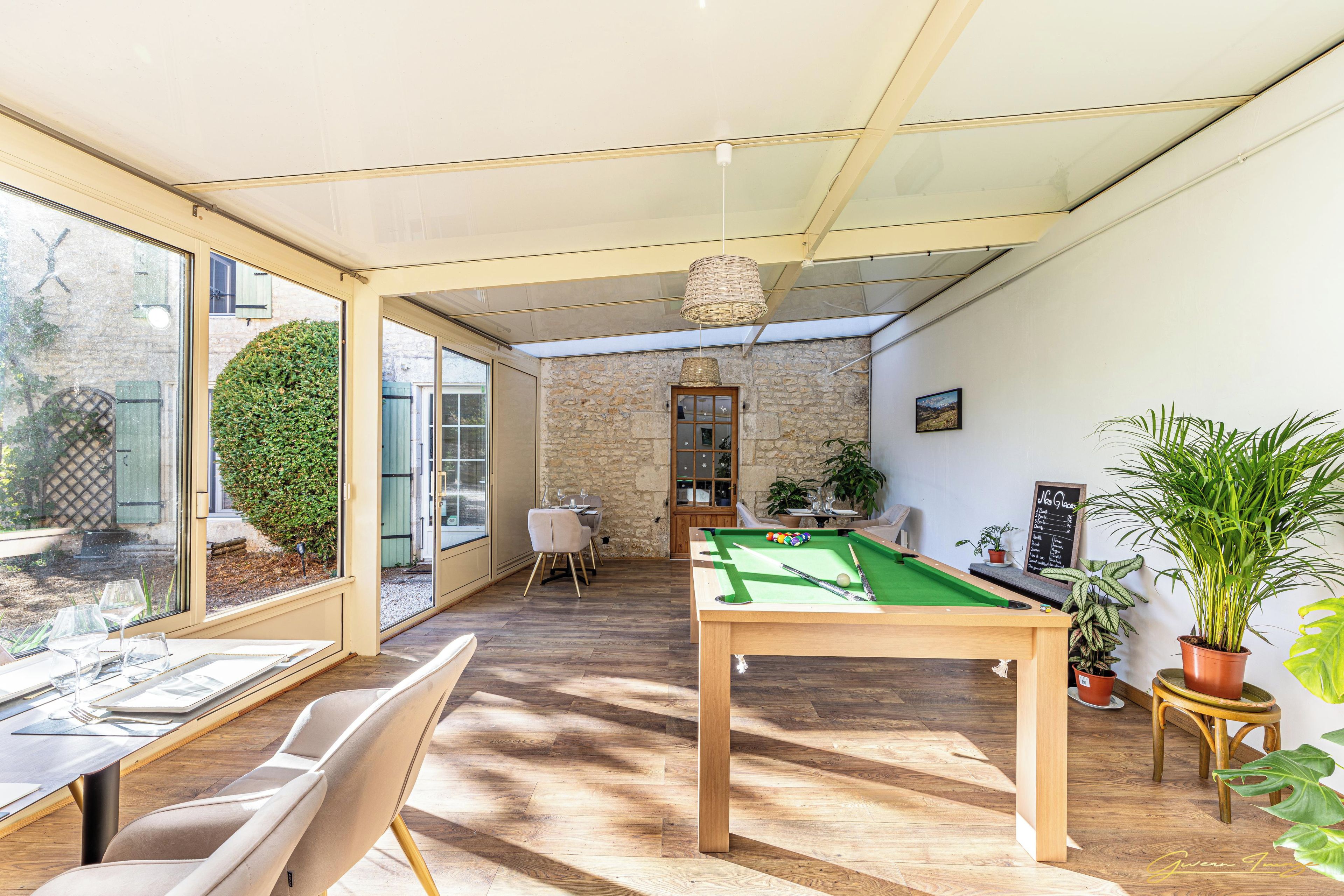 A room with a pool table, a dining area with chairs and a table, and a view through a glass door to an outdoor area with a hedge.