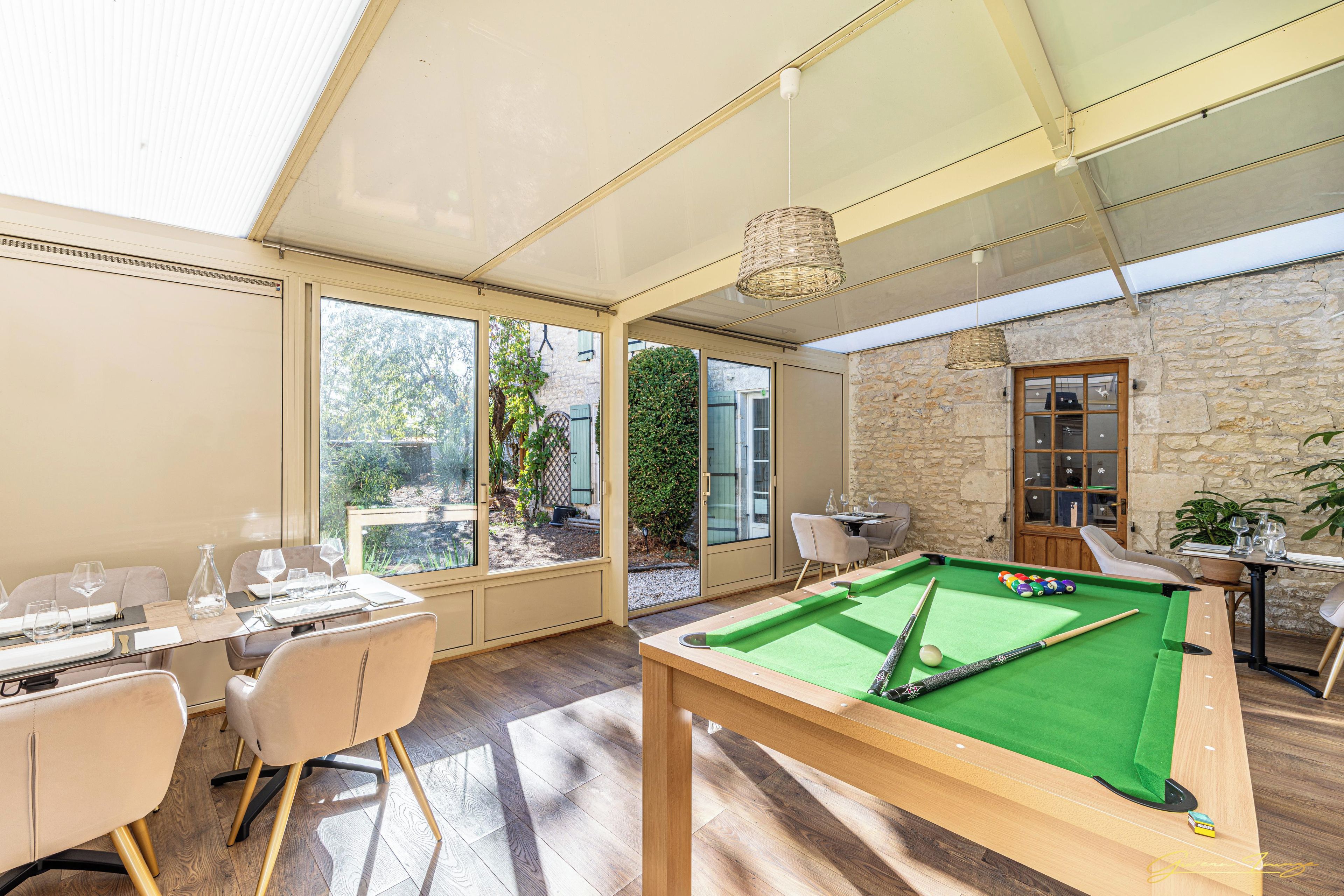 A pool table in a room with large windows, a dining area, and a stone wall.