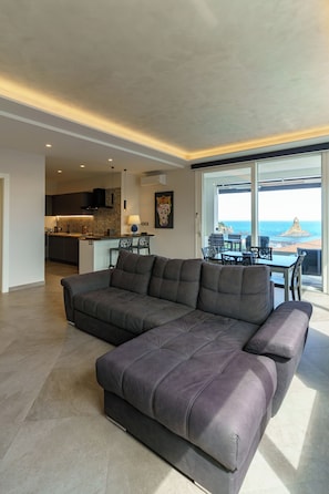 Una sala de estar moderna con un amplio sofá modular, una zona de comedor con mesa y sillas, y una cocina al fondo.