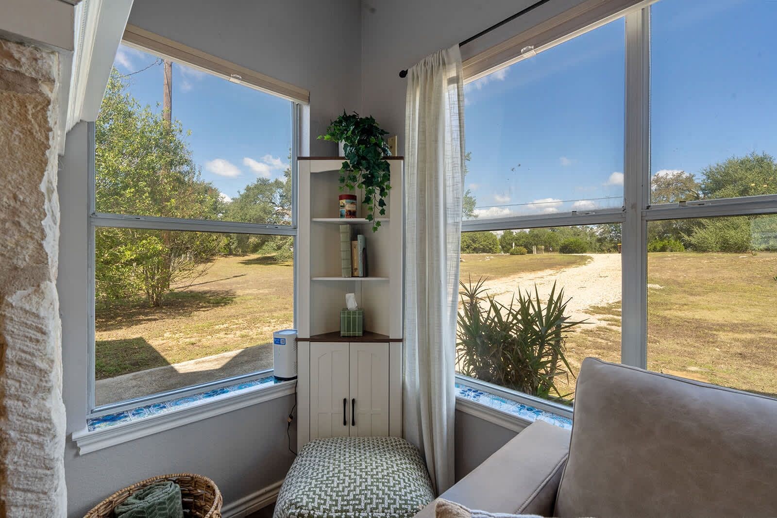 A room with a view of a grassy area, a window with a view, a shelf with items, and a cushion.