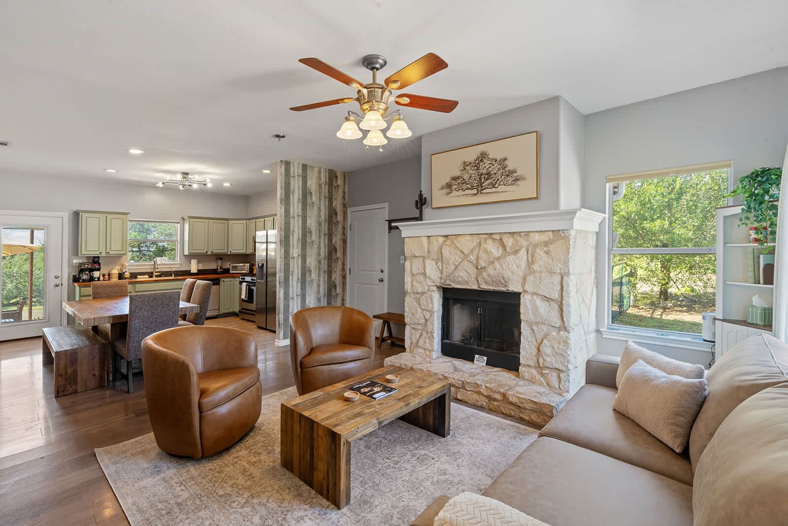 A modern living room with a fireplace, leather armchairs, a wooden coffee table, and a view of the kitchen and outdoor area.