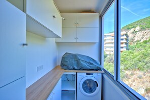 A compact laundry room with a washing machine, a drying rack, and a window with a view of a hillside. - Le Balcon des Sanguinaires - Vue sur la mer (Ajaccio)