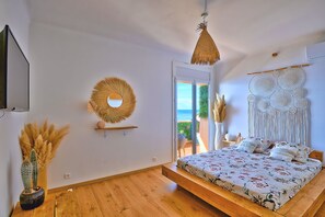 A bedroom with a bed, a wall-mounted TV, a decorative wall hanging, and a view of the outdoors. - Le Balcon des Sanguinaires - Vue sur la mer (Ajaccio)