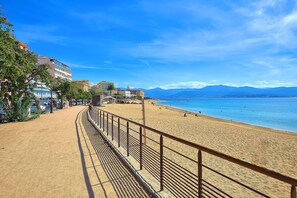 A coastal walkway with a metal railing, leading to a beach with clear blue water and mountains in the background. - Le Balcon des Sanguinaires - Vue sur la mer (Ajaccio)