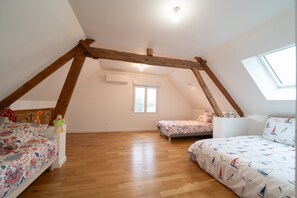 A bedroom with a sloped ceiling, wooden beams, a skylight, a bed with floral bedding, and a small table with toys. - Le Clos d'Adèle - Maison climatisée pour 8 (La Neuville-Bosmont)