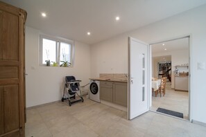 A modern kitchen with a washing machine, a dining area with a table and chairs, and a wooden cabinet. - Le Clos d'Adèle - Maison climatisée pour 8 (La Neuville-Bosmont)