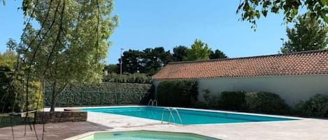 A swimming pool with a wooden deck, surrounded by greenery and a building with a tiled roof.