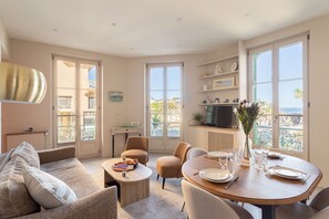 A well-lit living room with a sofa, chairs, a dining table set for two, and a television. - Le 50 Emeraude - 200m de la Plage (Dinard)