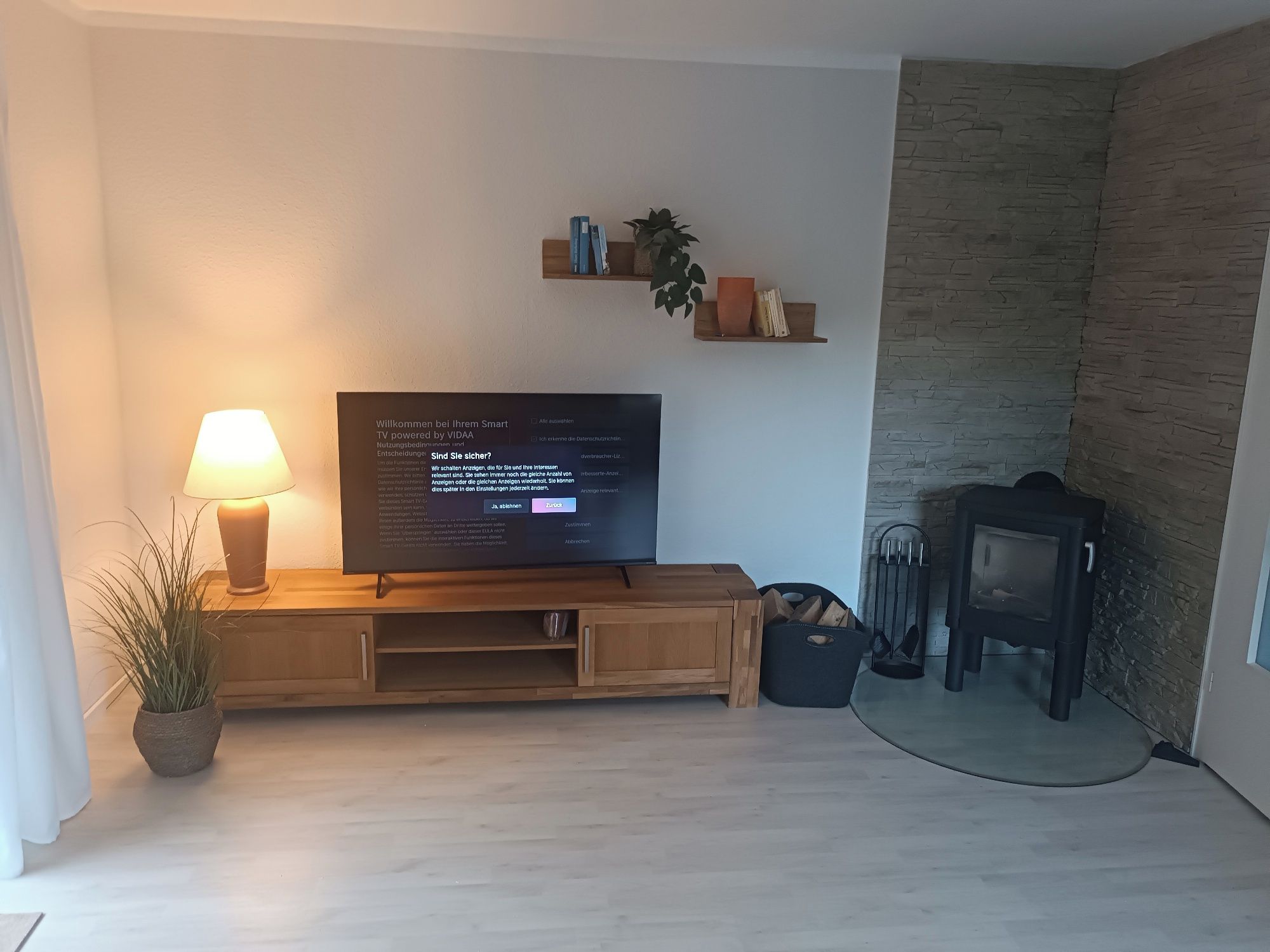 Ein modernes Wohnzimmer mit einem Flachbildfernseher auf einem Holzständer, einer Lampe, einem Topfpflanzen und einer Wand mit Steinoptik.