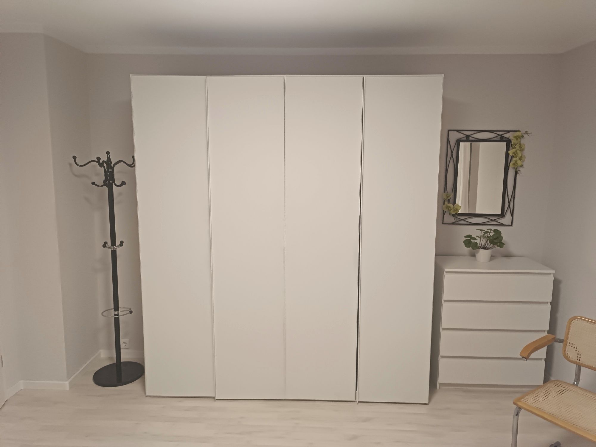 Ein Zimmer mit einem weißen Kleiderschrank, einem Wäscheständer, einem kleinen weißen Schminktisch, einem Stuhl und einem Spiegel mit einer Pflanze.