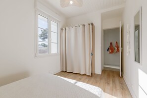 A bedroom with a large bed, a window, a door leading to a bathroom, and a hallway with a mirror and hanging bags. - Le Répit familial - Maison avec Jardin pour 6 (Saint-Jean-de-Monts)