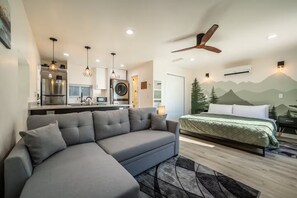 Ein modernes Schlafzimmer mit einem großen Bett, einem grauen Sofa, einem Deckenventilator und einer Küche im Hintergrund.