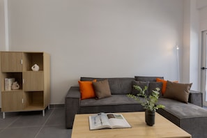 A modern living room with a grey sofa, a wooden cabinet, and a coffee table with a book and a vase of green leaves. - 3bdr Apartment in Kallithea (Kallithea)