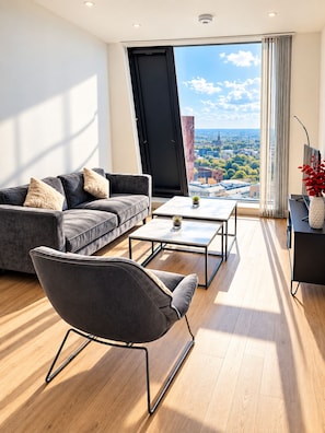 Ein modernes Wohnzimmer mit Sofa, Couchtisch und Sessel, ein großes Fenster mit Blick auf die Stadt und ein kleiner Tisch mit Pflanze.