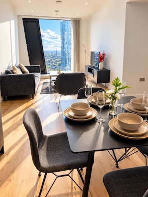 A modern dining area with a table set for four, a sofa, and a view of a cityscape through a large window. - Luxury 2-Bed Apartment in Manchester City Centre (Manchester)