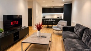 A modern living room with a flat-screen TV, a coffee table with a vase, a grey sofa, and a dining area in the background. - Luxury 2-Bed Apartment in Manchester City Centre (Manchester)