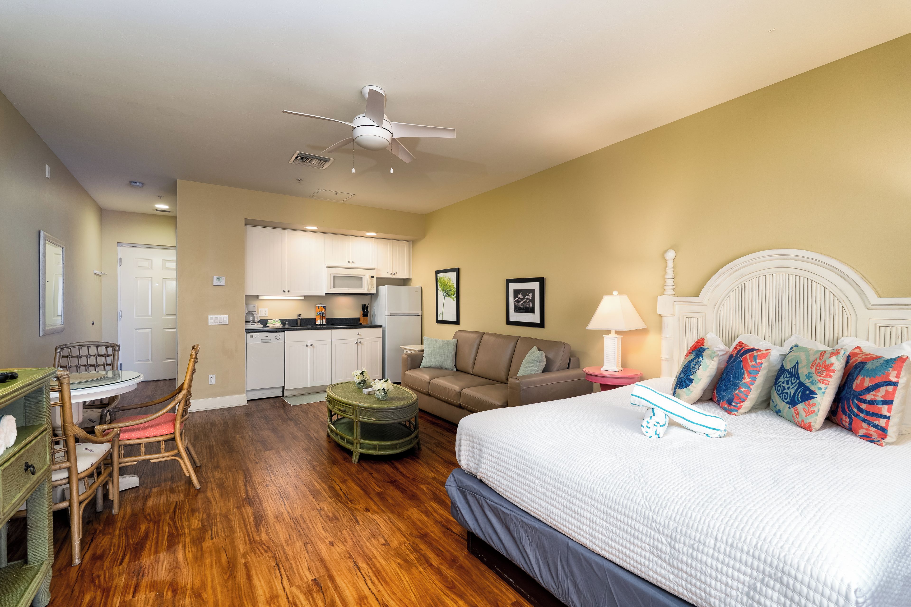 A bedroom with a bed, a dining table with chairs, a kitchen area, and a ceiling fan.