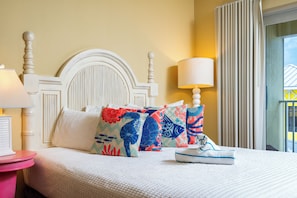 Een netjes opgemaakt bed met een wit hoofdeinde, meerdere kussens met kleurrijke patronen, een bedlampje en een raam met gordijnen.