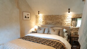 A bedroom with a stone wall, a bed with striped bedding, a nightstand with a lamp, and a framed picture on the wall. - The Little Cottage Kingham (Kingham)