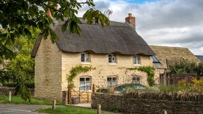 A stone house with a thatched roof, ivy-covered walls, and a stone wall in front. - The Little Cottage Kingham (Kingham)