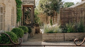 A stone building with a wooden archway leading to a garden area with various plants and a wooden fence. - The Little Cottage Kingham (Kingham)