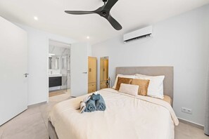 A modern bedroom with a bed, a ceiling fan, an air conditioning unit, and a bathroom visible through an open door. - Qasita Quracao - the Breeze Blue Bay Resort (Sint Michiel)