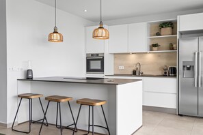 Cozinha moderna com ilha central, banquetas, iluminação pendente e eletrodomésticos embutidos.