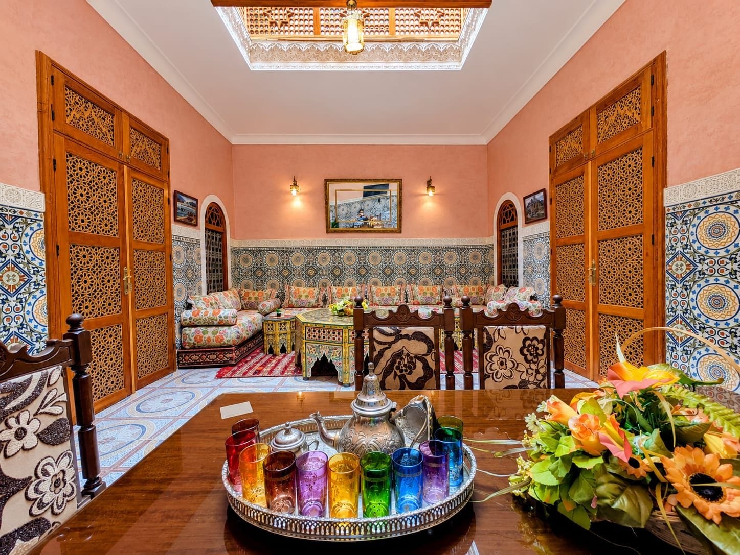 A traditional Moroccan room with a sofa, a dining table with a tray of colorful glasses, and a decorative wall.