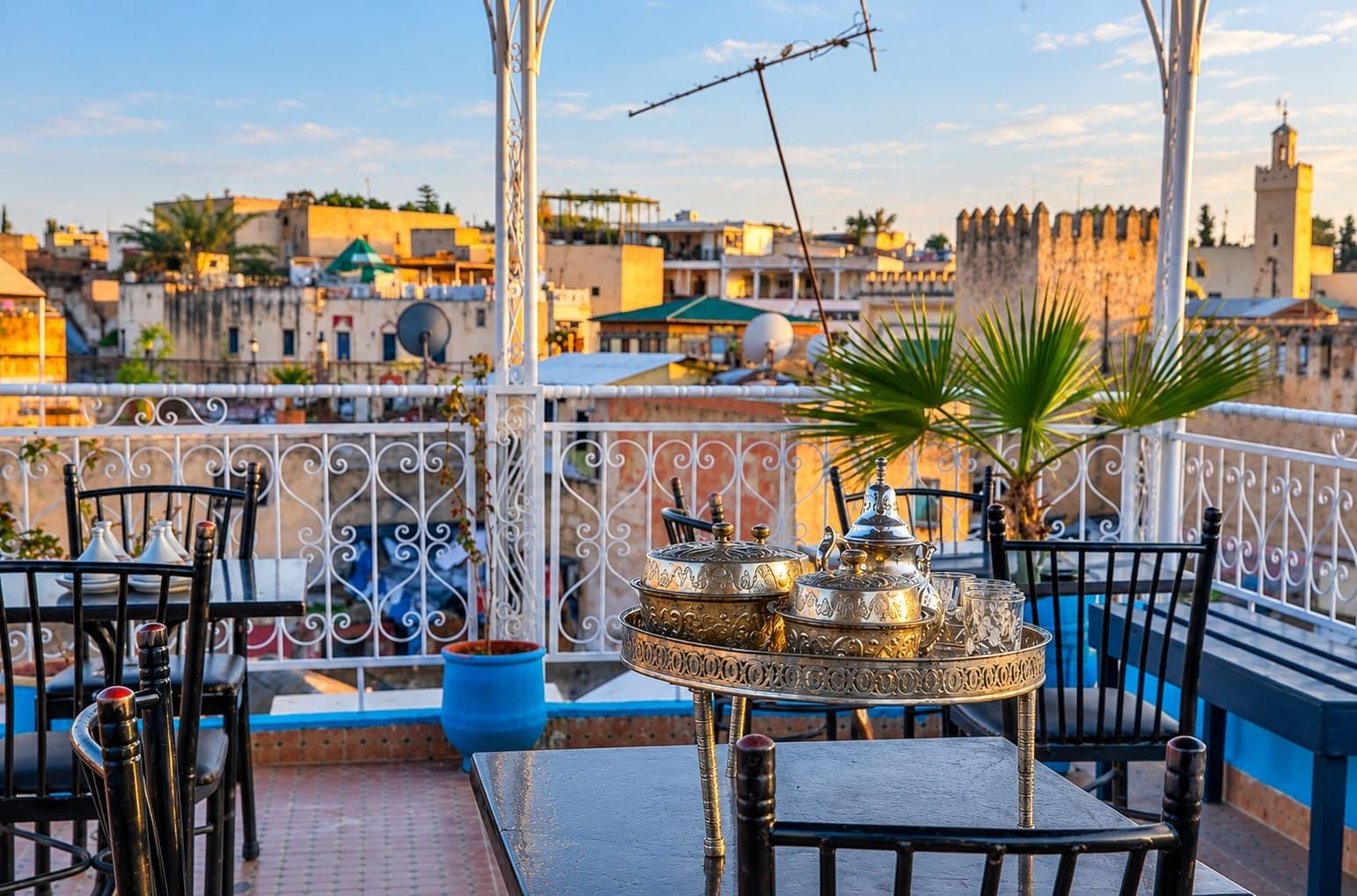A terrace with a table set for dining, surrounded by metal railings, and overlooking a cityscape with historic buildings.