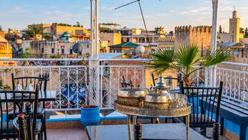A terrace with a table set for dining, surrounded by metal railings, and overlooking a cityscape with historic buildings.