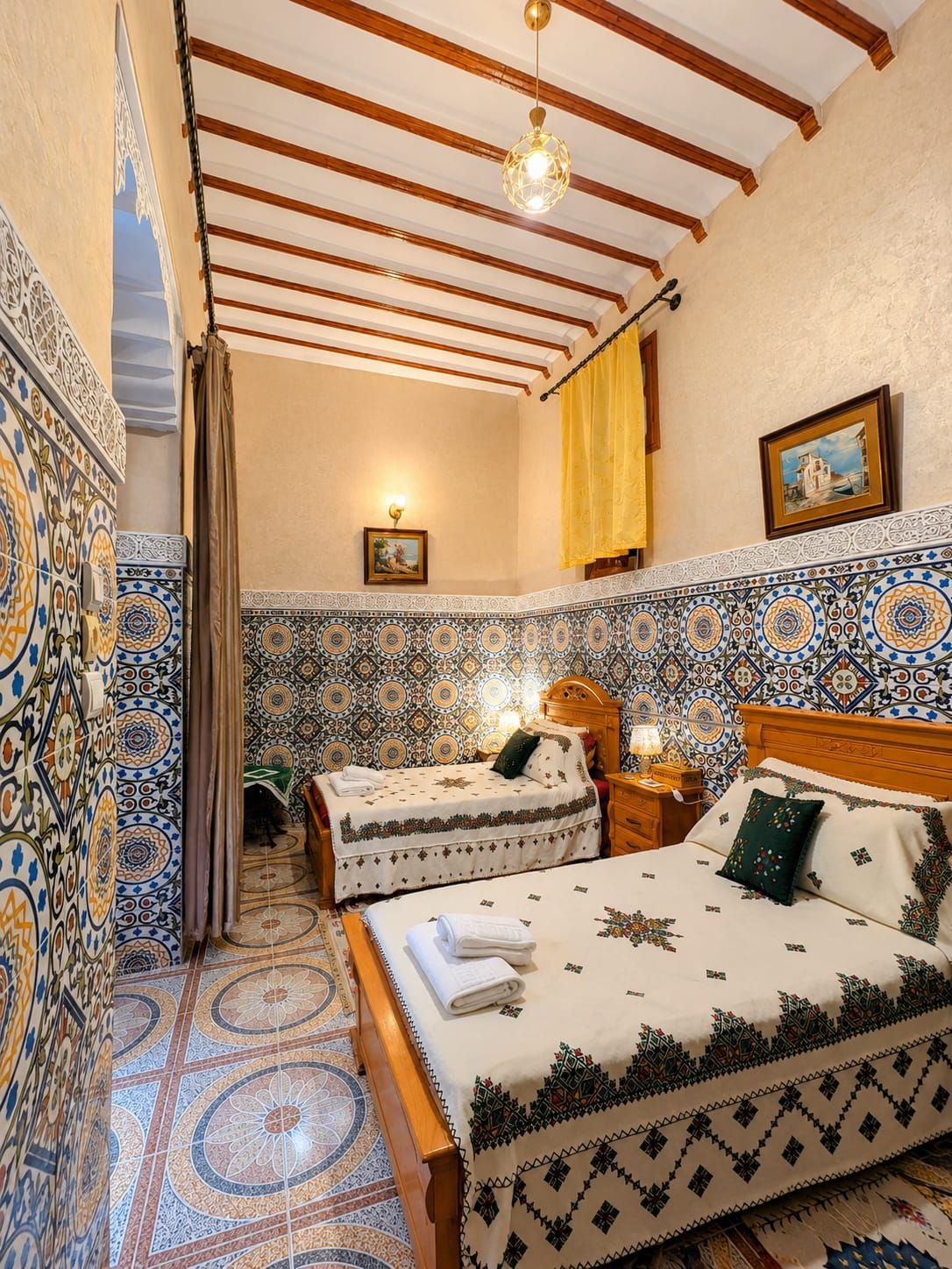 A room with two beds, patterned walls, a wooden ceiling, and a small table with a lamp.