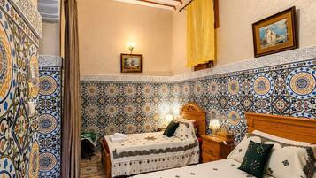 A room with two beds, patterned walls, a wooden ceiling, and a small table with a lamp.