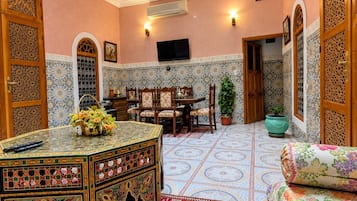 Una habitación con techo alto, televisor, zona de comedor con sillas y una alfombra decorativa.