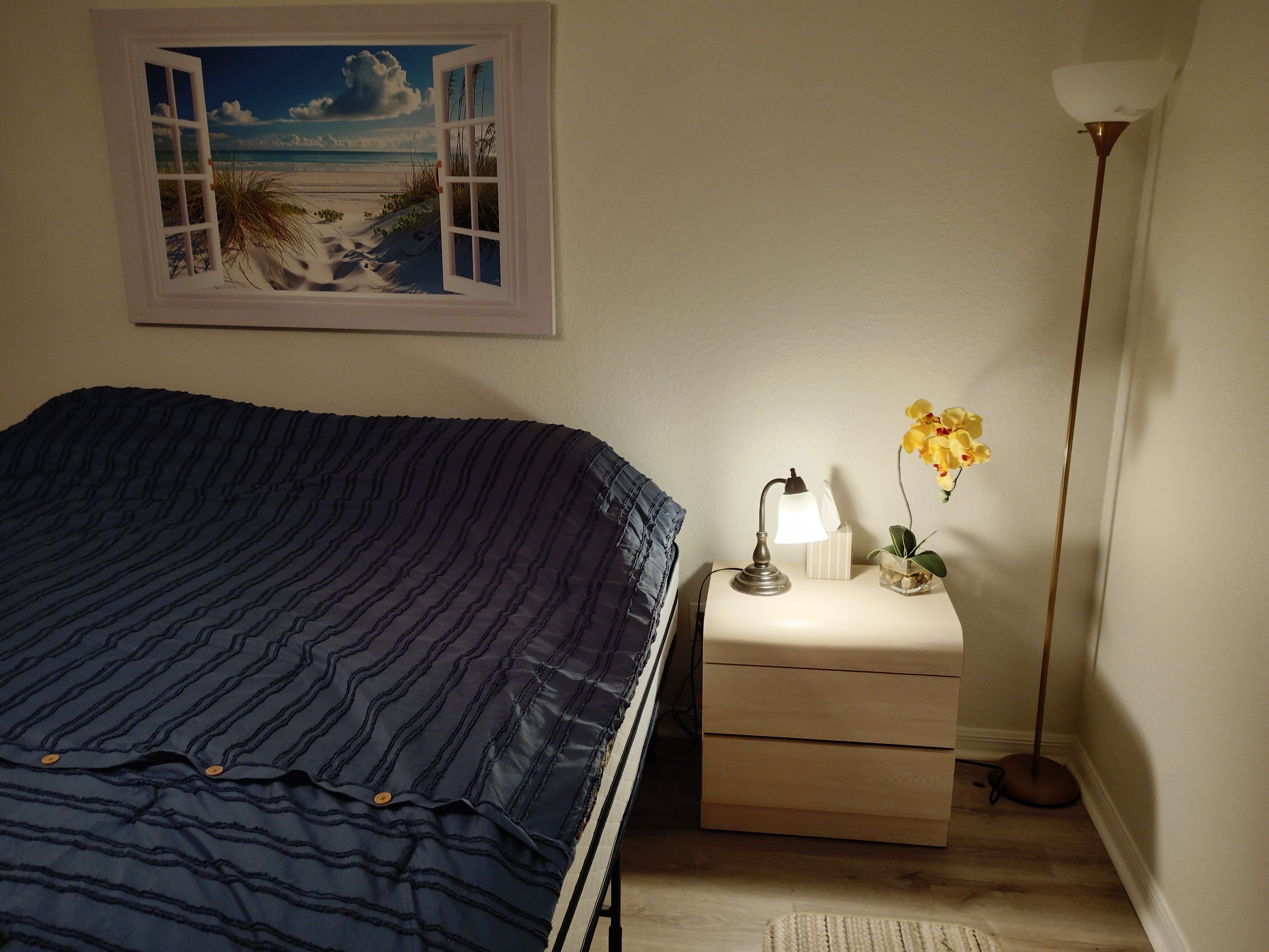 A bedroom with a bed, a nightstand with a lamp, a vase with flowers, and a painting of a beach scene.