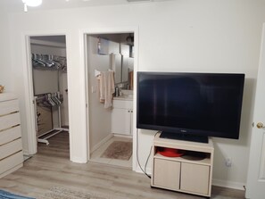 Ein Zimmer mit einem Flachbildfernseher auf einem Tisch, Holzboden, einem Schrank mit Kleidern und einer Tür zu einem anderen Raum.
