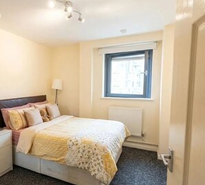 Ein Schlafzimmer mit Bett, Nachttisch, Lampe, Fenster und Heizkörper.