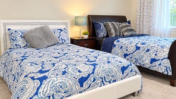 Un dormitorio con dos camas, cada una con ropa de cama estampada en azul y blanco, una mesita de noche con lámpara y una ventana con cortinas.