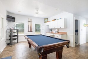 A pool table in a room with white walls, a television mounted on the wall, and a window with a view of greenery. - Corolla Retreat w/ Pool, Hot Tub & Game Rm (Corolla)