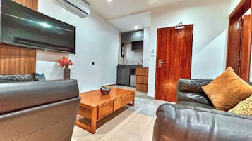 A modern living room with a flat-screen TV, leather sofas, a wooden coffee table, and a kitchen in the background.