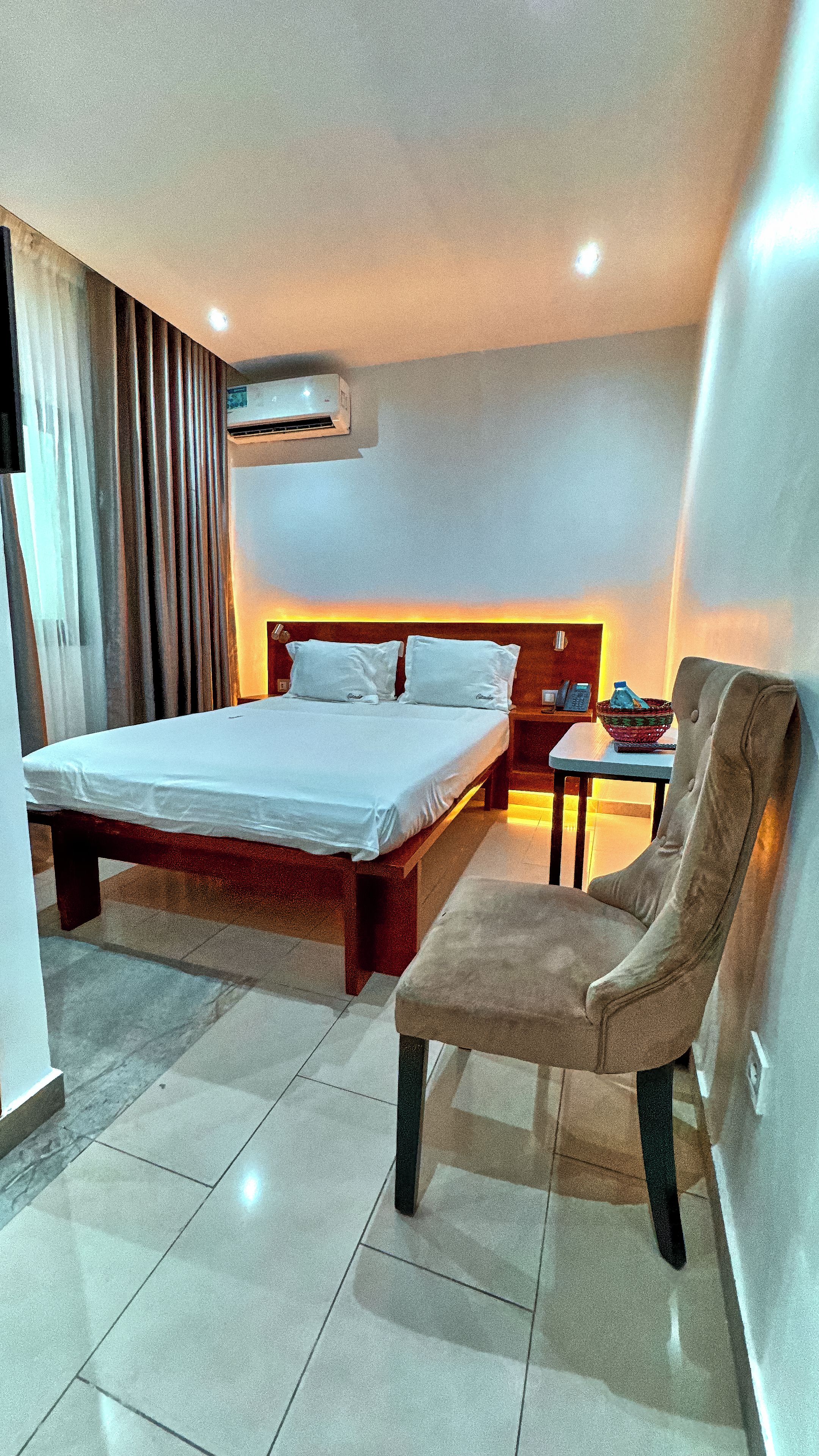 A hotel room with a bed, a chair, a table, an air conditioning unit, and a nightstand.