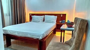 A hotel room with a bed, a chair, a table, an air conditioning unit, and a nightstand.
