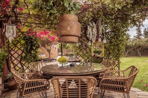 A round glass table surrounded by wicker chairs in an outdoor garden setting with a pergola overhead. - Filoxenia Luxury Studios & Apartments (Zakynthos)