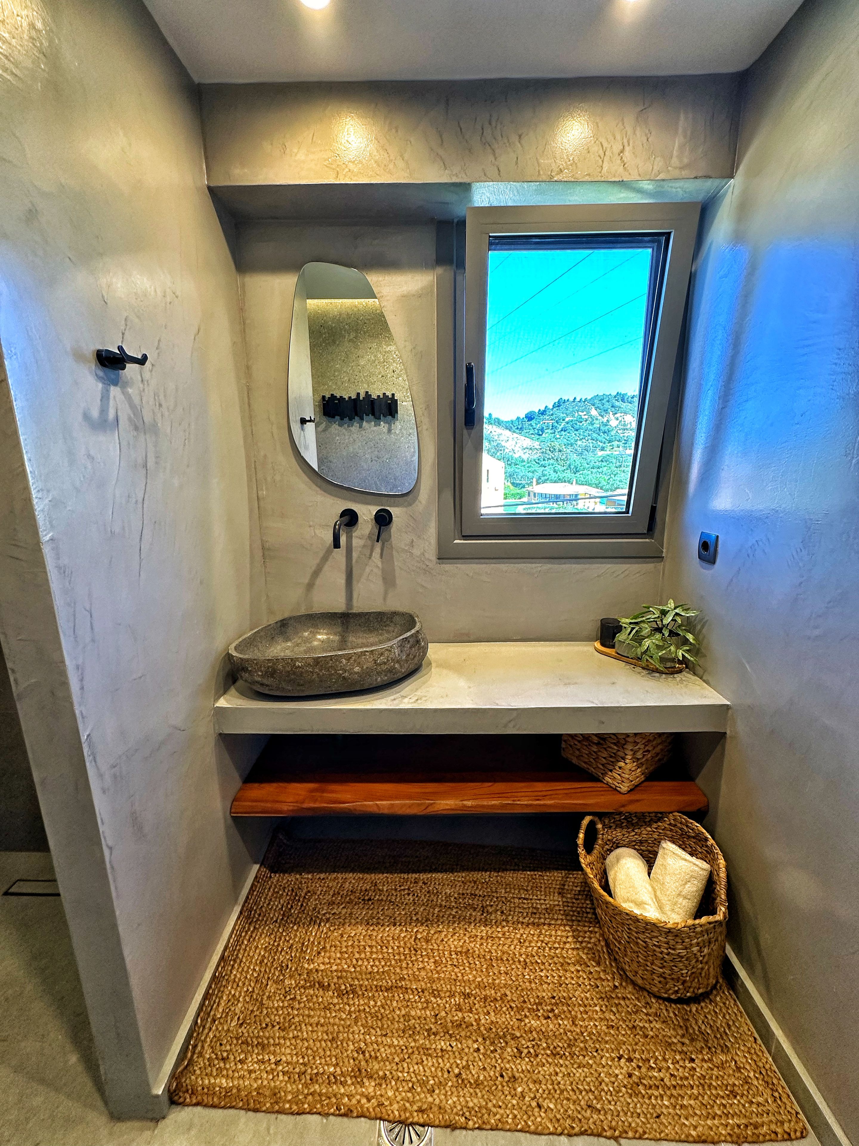 Un baño con un lavabo de piedra, un espejo, una ventana con vistas y un tapete tejido en el suelo.