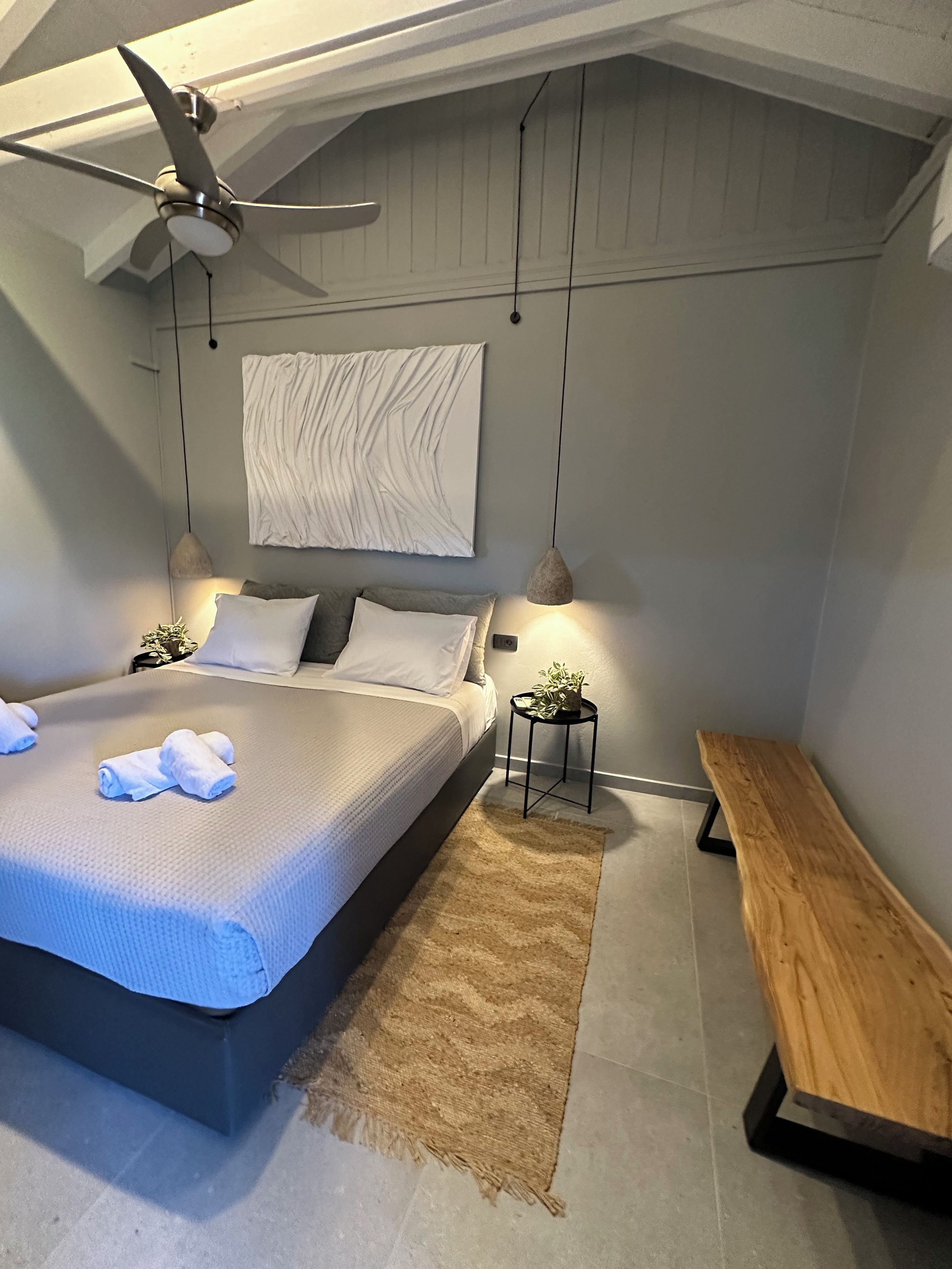 Un dormitorio moderno con cama, mesitas de noche, un banco, ventilador de techo e iluminación en la pared.