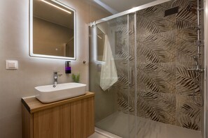 A modern bathroom with a glass shower enclosure, a white sink, and a mirror above it. - Minimal City View Apartment (Kallithea)